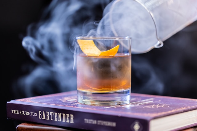 A cocktail glass, filled with a whiskey-based drink garnished with a citrus peel, sits atop the book "The Curious Bartender."