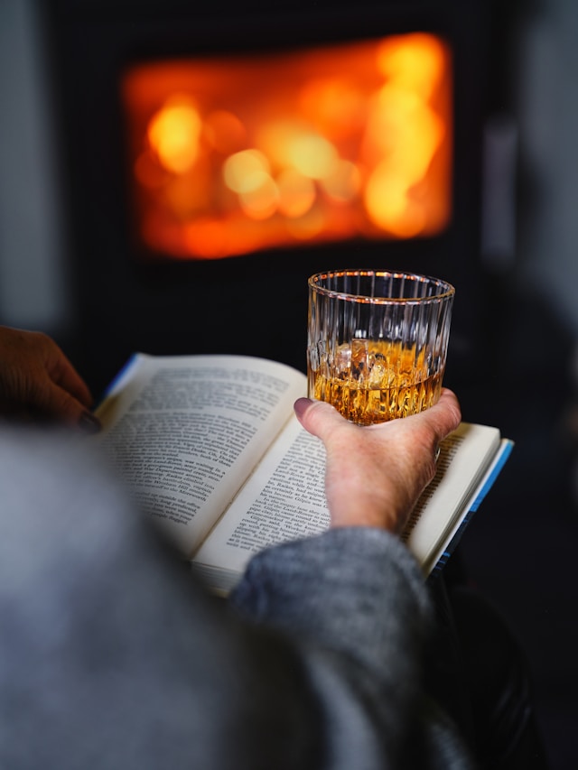 A person holds a glass of whiskey in one hand while reading a book, with the warm glow of a fire in the background.