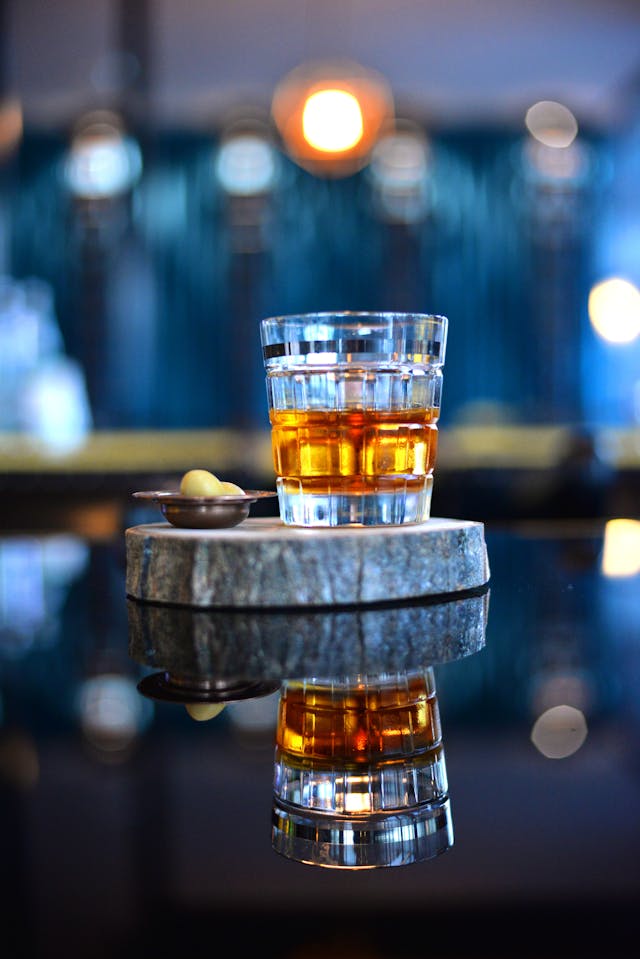 A glass of whisky and olives on a wooden tray, reflecting on a shiny surface.