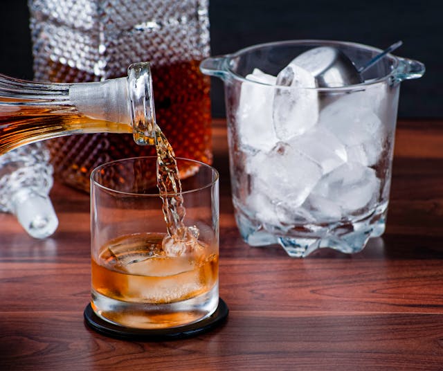 A glass of whiskey being poured from a decanter, with an ice bucket on the side.
