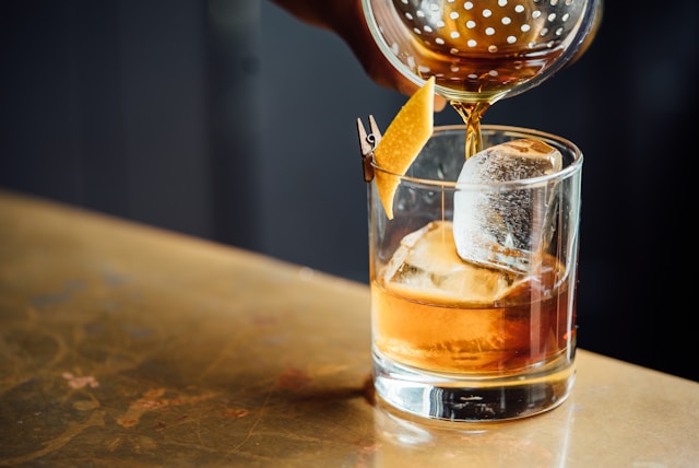 A poured glass of blended whiskey served over large ice cubes, garnished with a twist of citrus peel on a table.