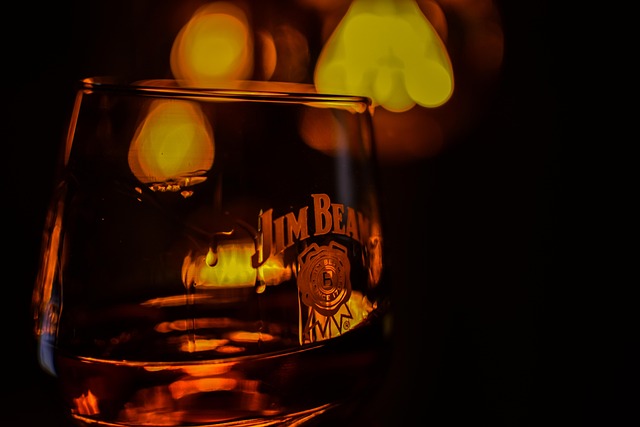 A close-up of a Jim Beam branded glass with whisky, illuminated by warm, blurry lights in the background.