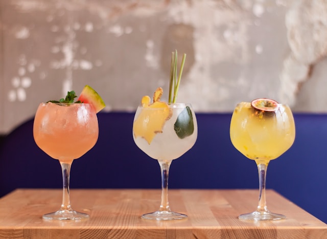 Three cocktails in glasses garnished with fresh ingredients like watermelon, mint, ginger, lemongrass, and passion fruit. 