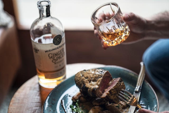 A person holding a glass of whisky near a plate of steak alongside a bottle of Misunderstood Ginger Spiced Whiskey.