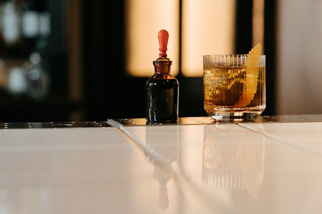 A glass of rye whiskey with a garnish and a dropper bottle of bitters on a bar counter.