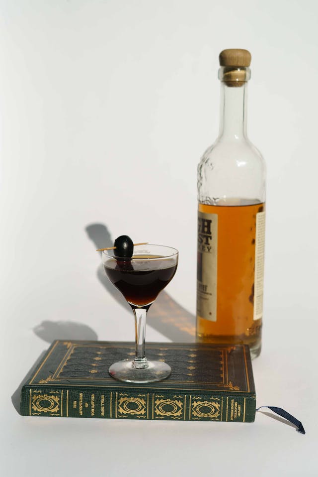 Manhattan cocktail garnished with a cherry on top of a vintage book, with a whiskey bottle beside.