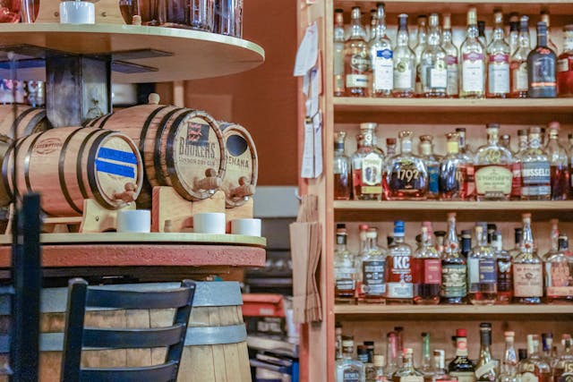 Wooden barrels of spirits on display in a bar, with shelves of whiskey and liquor bottles in the background.