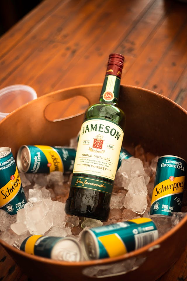 A bottle of Jameson Irish Whiskey and cans of Schweppes lemonade sit in a bucket filled with ice, placed on a wooden table.