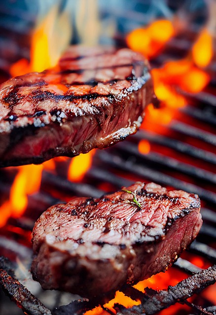 Two thick steaks sizzling on a hot grill over open flames, with visible grill marks and charred edges.