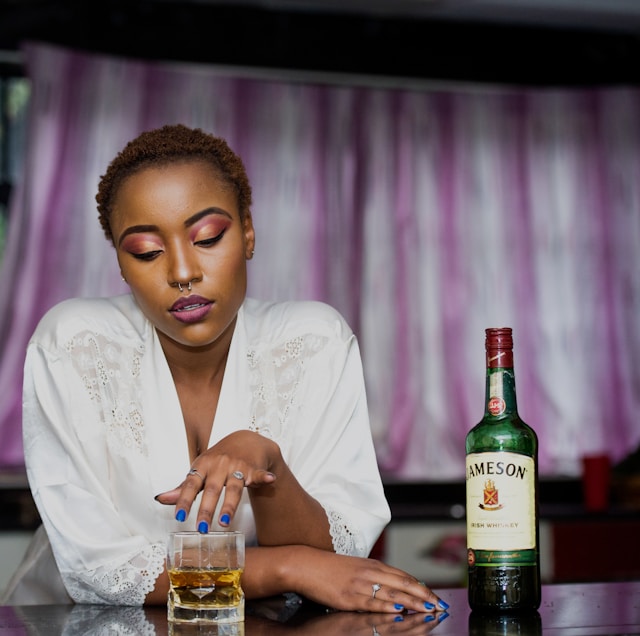 A woman with colorful makeup sits at a table with a glass of whiskey and a bottle of Jameson Irish Whiskey.