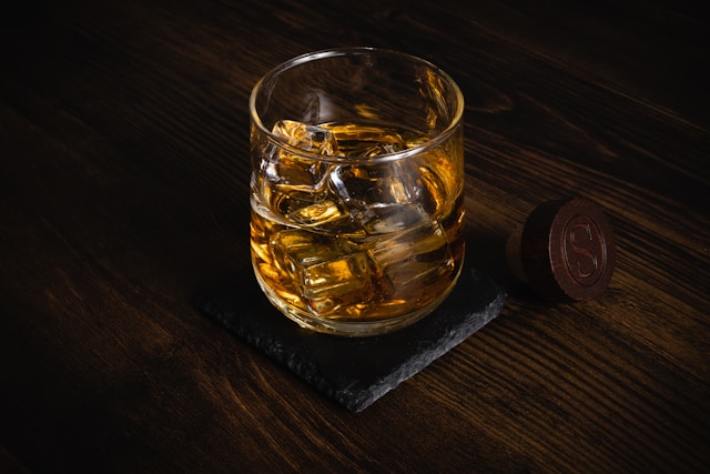 A glass of bourbon on the rocks rests on a stone coaster atop a rustic wooden table, exuding a moody ambiance.