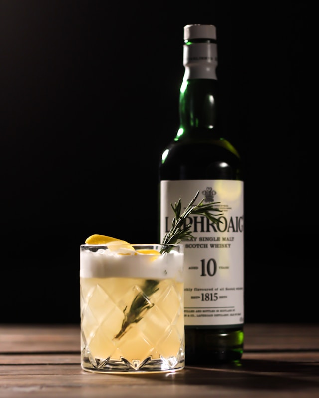 A Laphroaig 10 whisky bottle stands behind a whiskey sour cocktail garnished with rosemary and lemon peel.