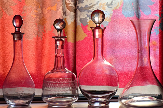 Four elegant whiskey decanters with decorative stoppers are arranged in front of a pink and gray floral-patterned background. 