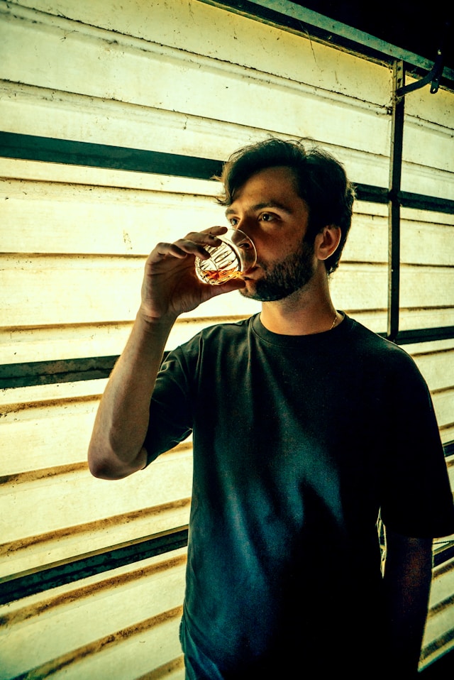 A man drinking whisky from a glass in front of a rustic window backdrop.