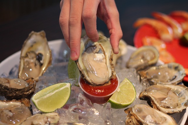 A hand holding an oyster over a cup of sauce, with limes and oysters on ice in the background.