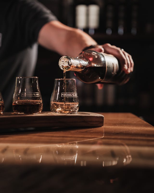 A person pours whiskey from a bottle into tasting glasses on a wooden board.