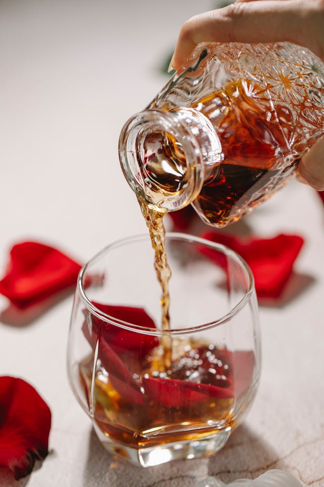 A close-up of a person pouring whisky from a decorative glass decanter into a tumbler glass that contains red rose petals. 