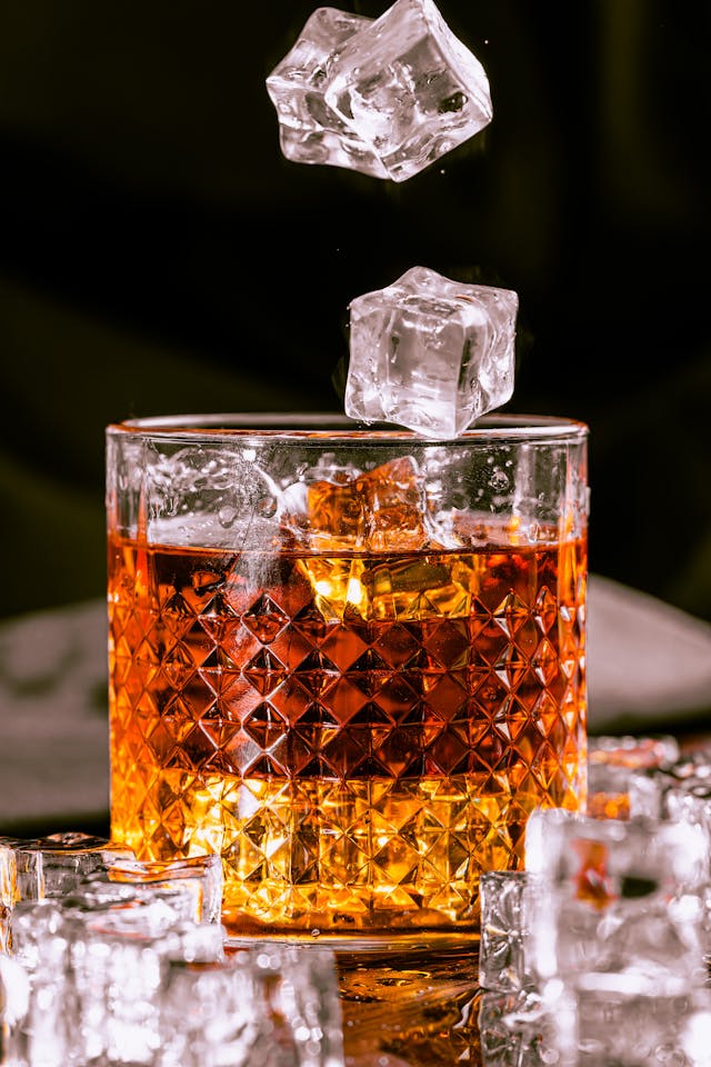 A close-up of a glass of whiskey with ice cubes being dropped into it, surrounded by scattered ice on a reflective surface.