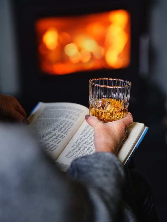 A person holding a glass of whisky in one hand while reading a book in the other.