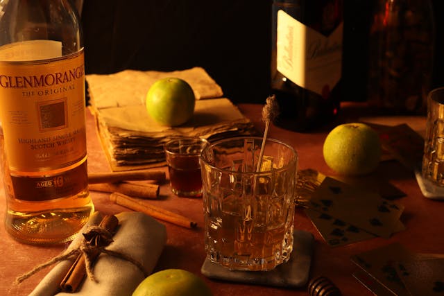 A bottle of Glenmorangie sits on a rustic table with green apples, cinnamon sticks, and a glass of whisky.