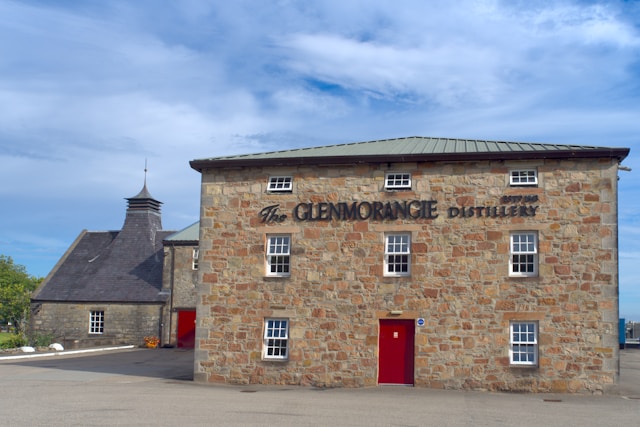 The front of the Glenmorangie Distillery, a traditional stone building with a red door and sign, under a clear blue sky.