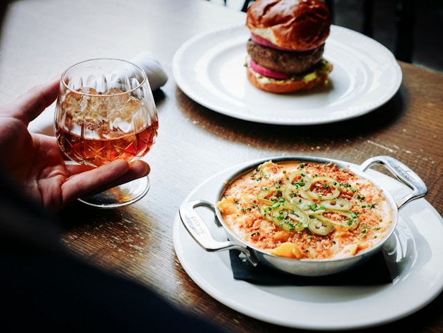 A glass of whiskey, a dish of gourmet mac and cheese garnished with jalapeños, and a juicy burger in the background.