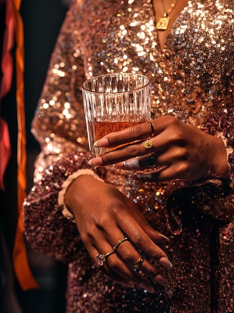 A close-up of a woman in a sparkling dress holding a whiskey glass, adorned with elegant rings, capturing a glamorous moment.