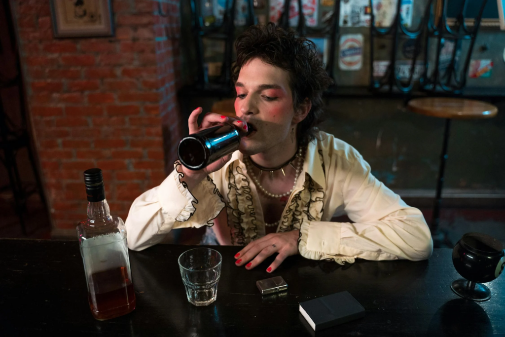 A person drinks from a beer bottle at a bar, with a whiskey bottle, glass, and lighter on the table in a rustic setting.