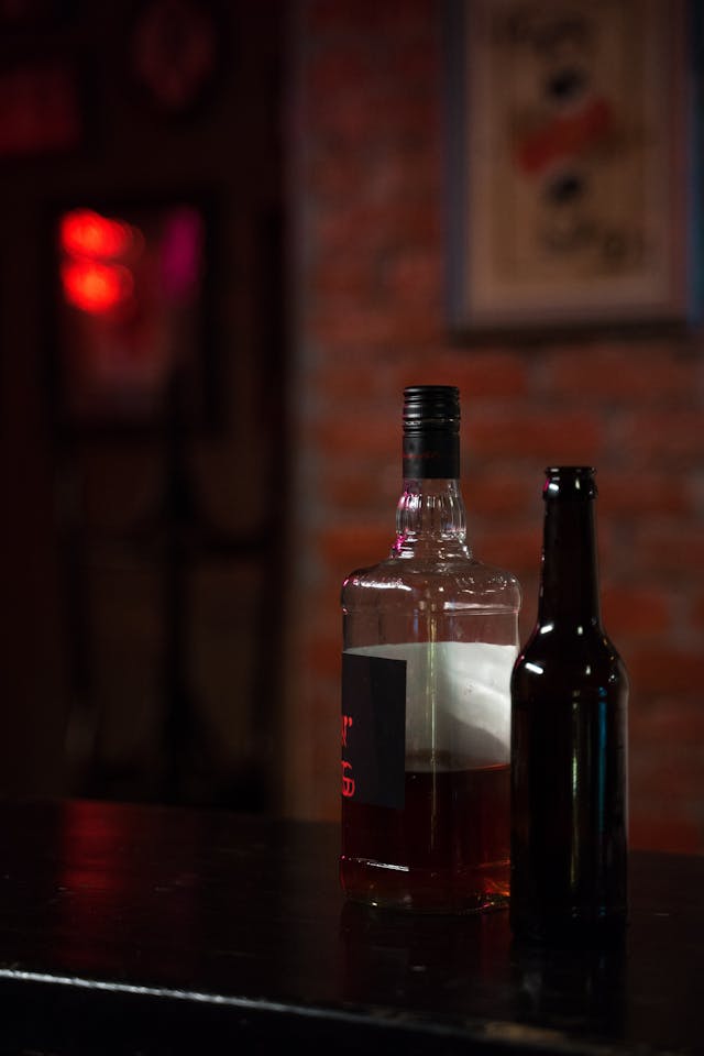 A whiskey bottle and a beer bottle side by side on a dark surface with a blurred brick background.