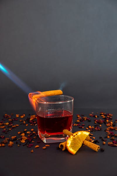 A flaming whiskey cocktail in a glass, garnished with a lit cinnamon stick, surrounded by dried fruit and spices.