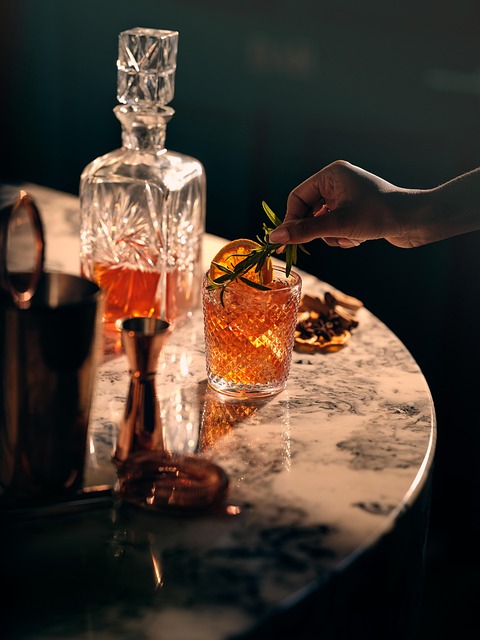 A hand garnishing an elegant whiskey cocktail with rosemary and orange on a marble counter.