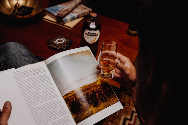 A person with a glass of corn whiskey while reading a book, with a bottle of "Corn Trooper" whiskey and cigars.