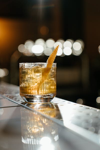 An elegant glass of whiskey with ice and a citrus twist on a reflective bar counter, with blurred lights in the background.