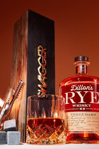 A bottle of Dillon’s Rye Whiskey with a glass, whiskey stones, and tongs, set against an orange background with light trails.