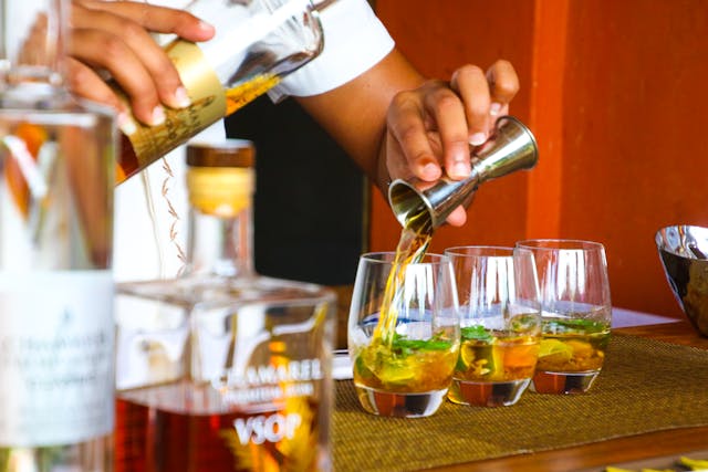 A bartender pouring whiskey into glasses with mint and lime, preparing cocktails at a bar with bottles in the foreground.